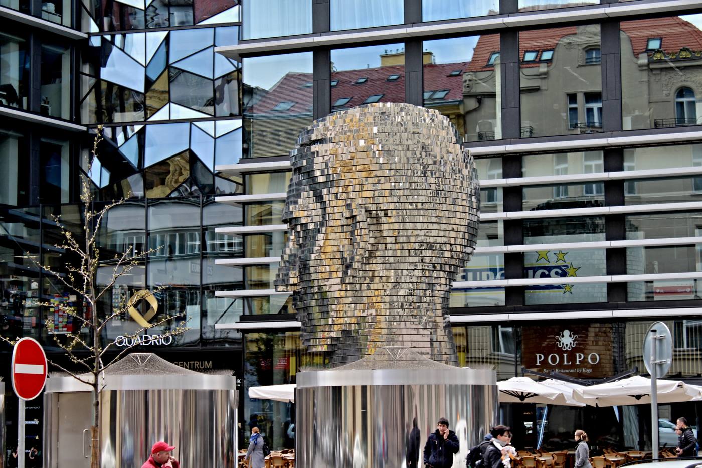 The Kafka head rising above café umbrellas in the square behind Quadrio