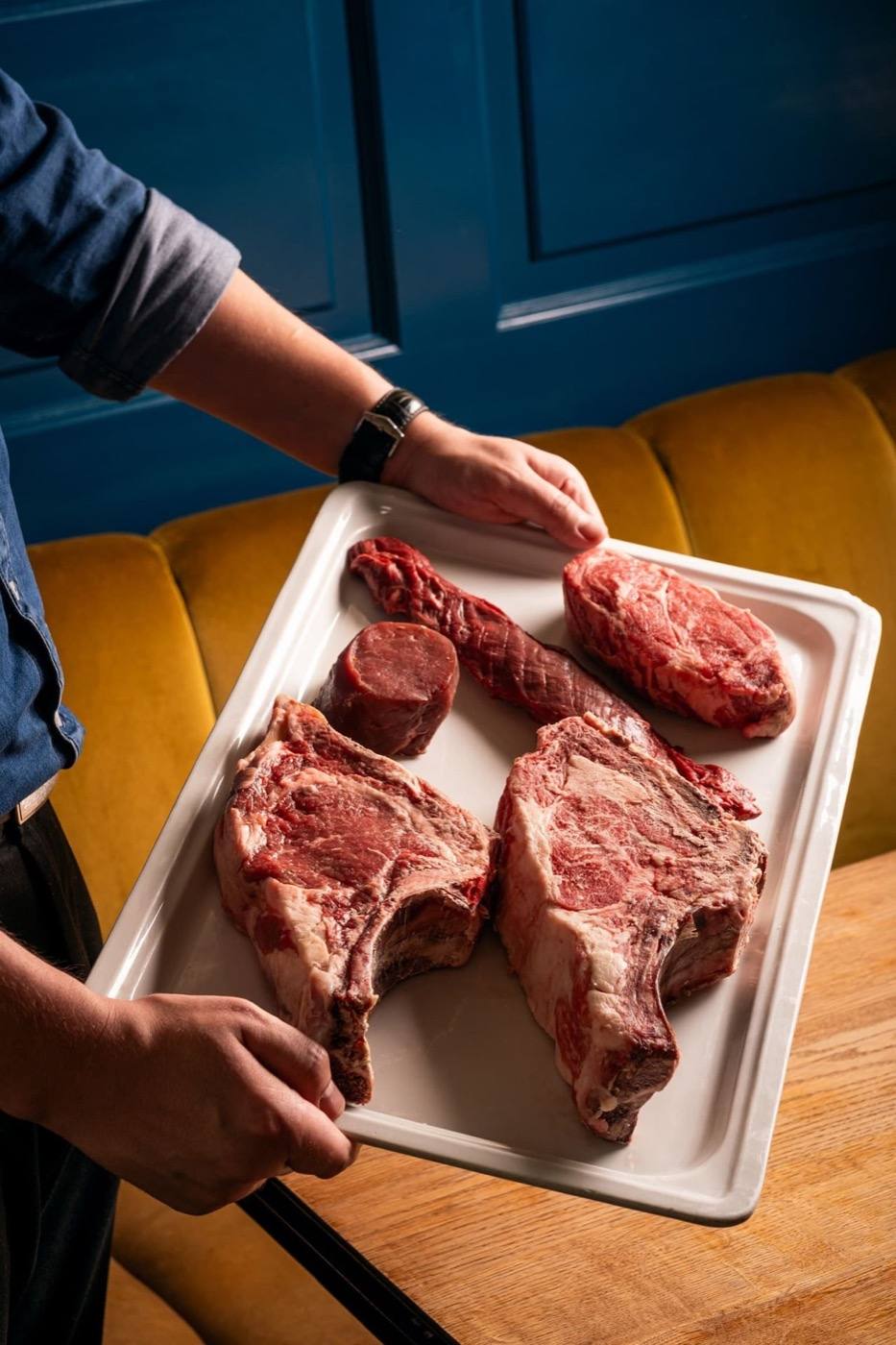 A waiter presenting a tray of raw dry-aged steaks, including a T-bone, for the charcoal grill