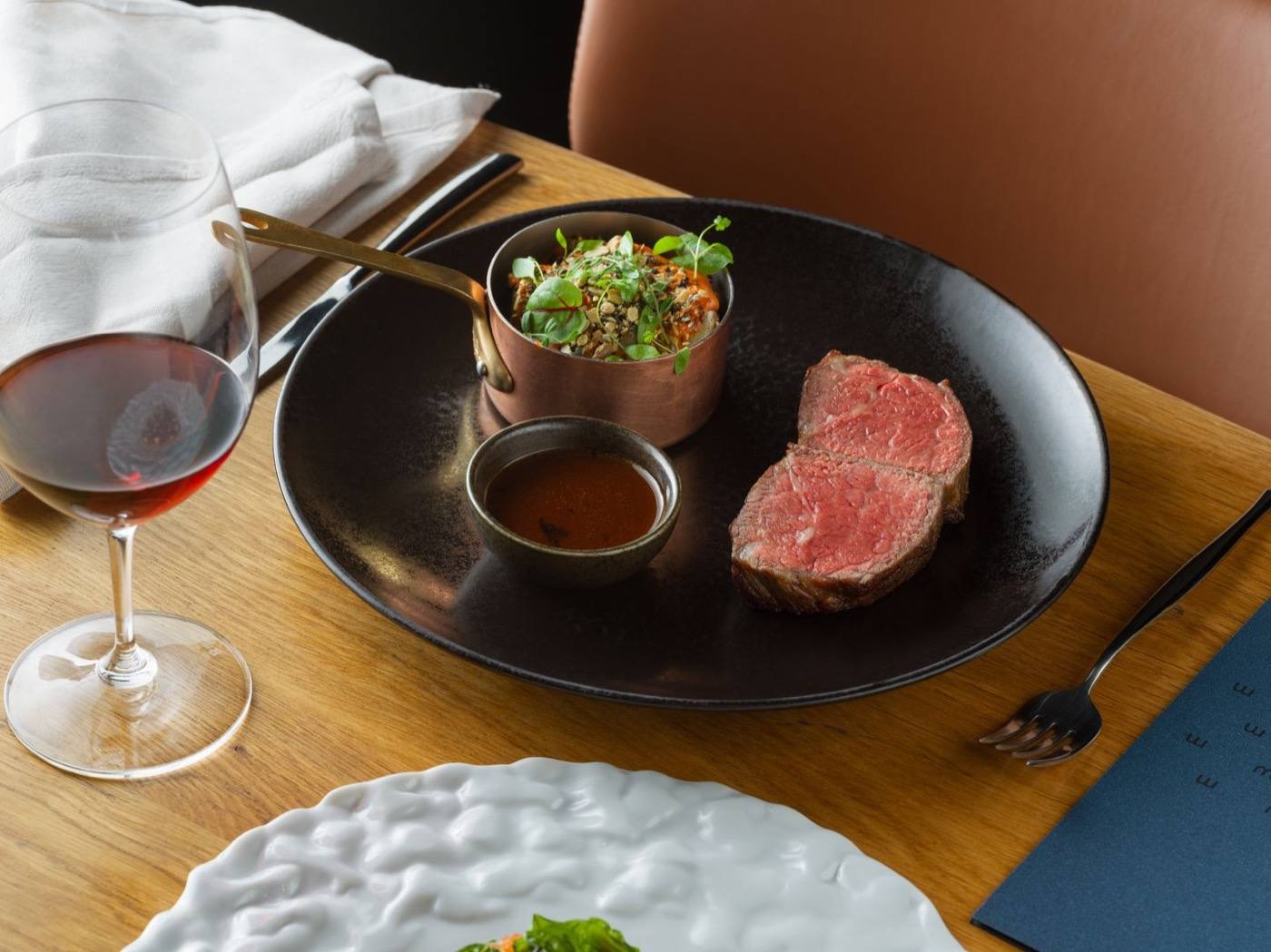Sliced beef fillet with a copper pot of salad and sauce, next to a glass of red wine