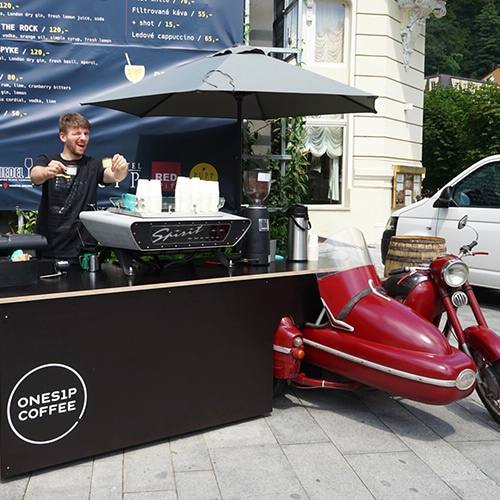 Onesip barista serving coffee from the pop-up bar next to a red vintage motorcycle with sidecar